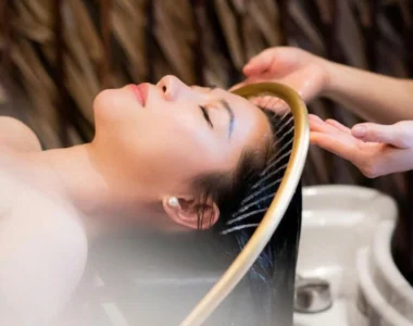 A woman lies with her eyes closed, receiving a soothing hair spa treatment from a therapist. Her hair is wet and being massaged, indicating a relaxing salon or spa experience.