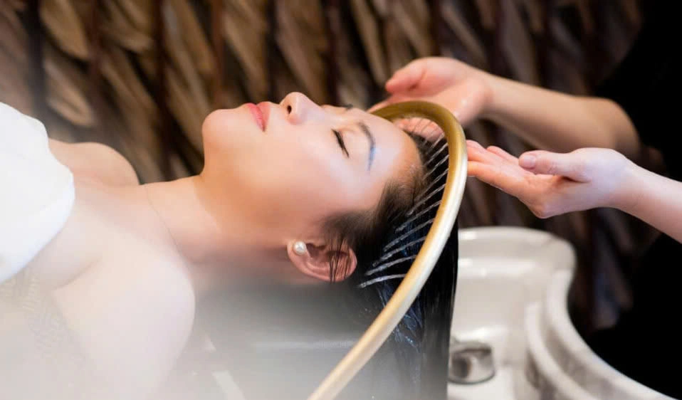 A woman lies with her eyes closed, receiving a soothing hair spa treatment from a therapist. Her hair is wet and being massaged, indicating a relaxing salon or spa experience.