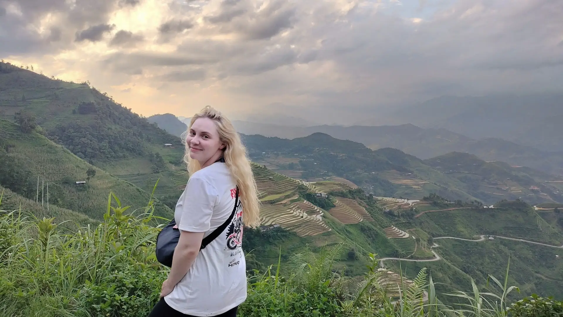 A smiling female traveler standing at a high viewpoint overlooking vast terraced rice fields and mountains in Ha Giang under dramatic clouds.