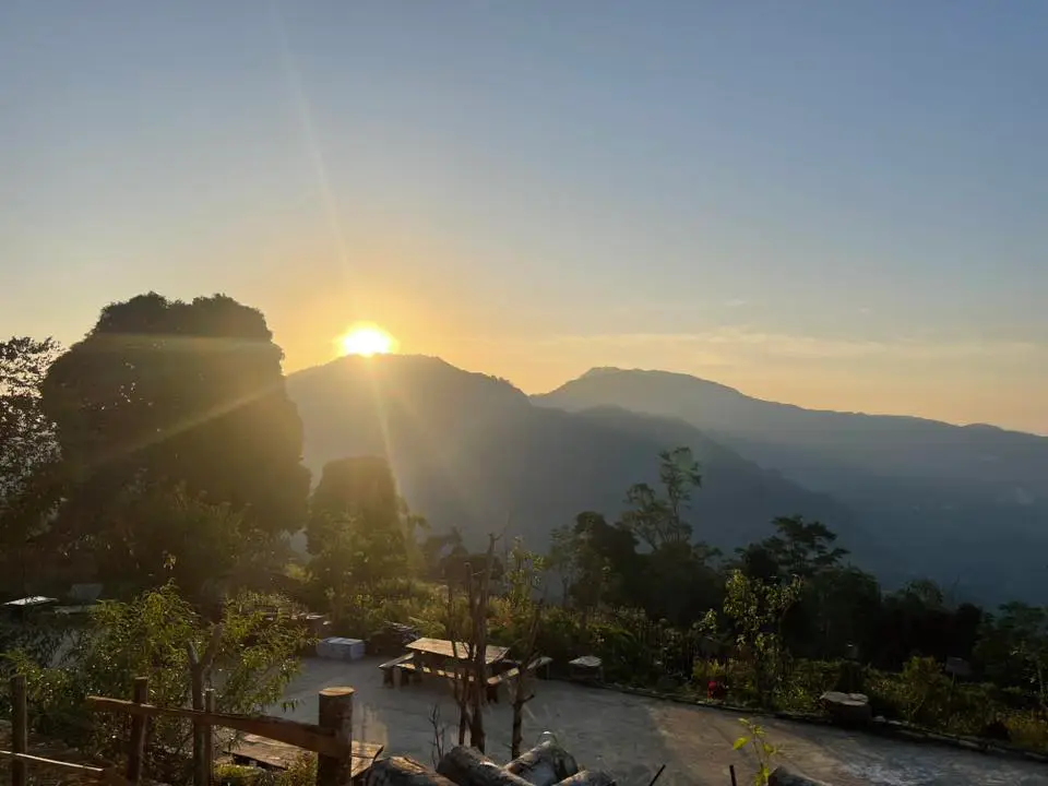 The sun rising over a distant mountain ridge, casting golden light over a rustic viewing platform with wooden tables in Ha Giang.