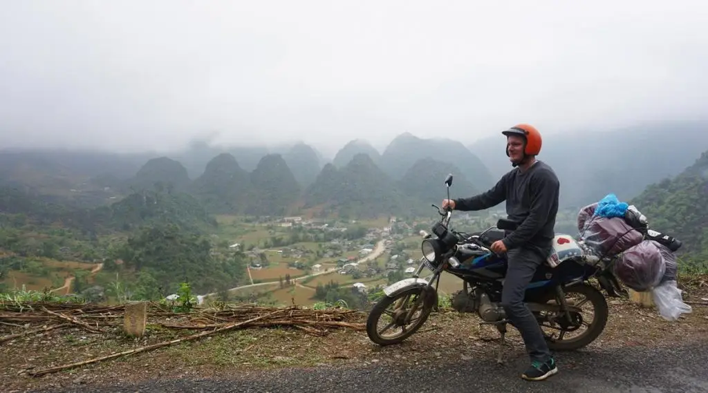 A traveler on a motorbike loaded with luggage, paused at a viewpoint overlooking a misty valley filled with conical karst mountains in Ha Giang.