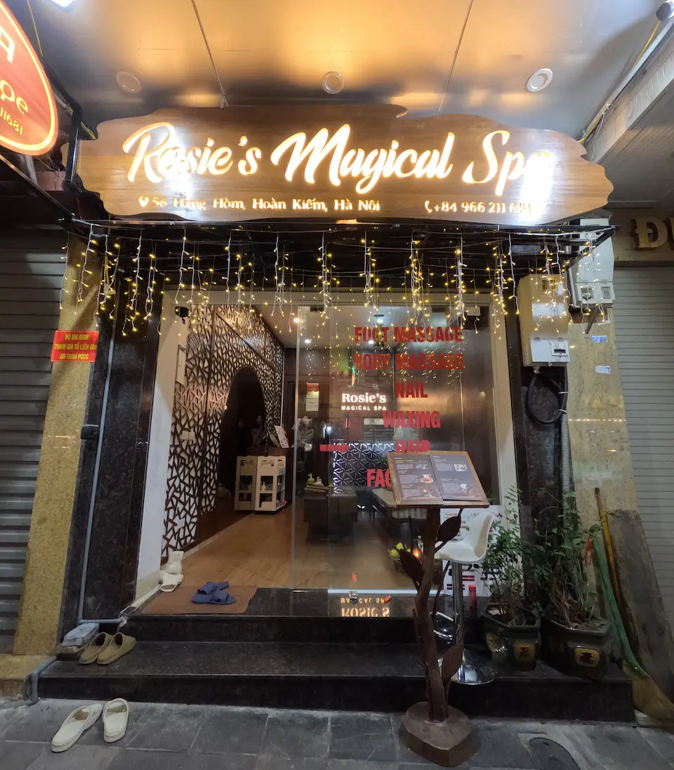 The illuminated storefront of Rosie's Magical Spa at 56 Hang Hom, Hanoi Old Quarter at night, featuring a wooden sign and fairy lights.