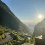 A dramatic winding asphalt road of the Ha Giang Loop cutting through steep, lush green mountains under a bright sun late in the day.