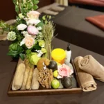 A wooden tray displaying fresh herbs, cinnamon sticks, lime, herbal compress balls, and rolled towels for spa treatments.